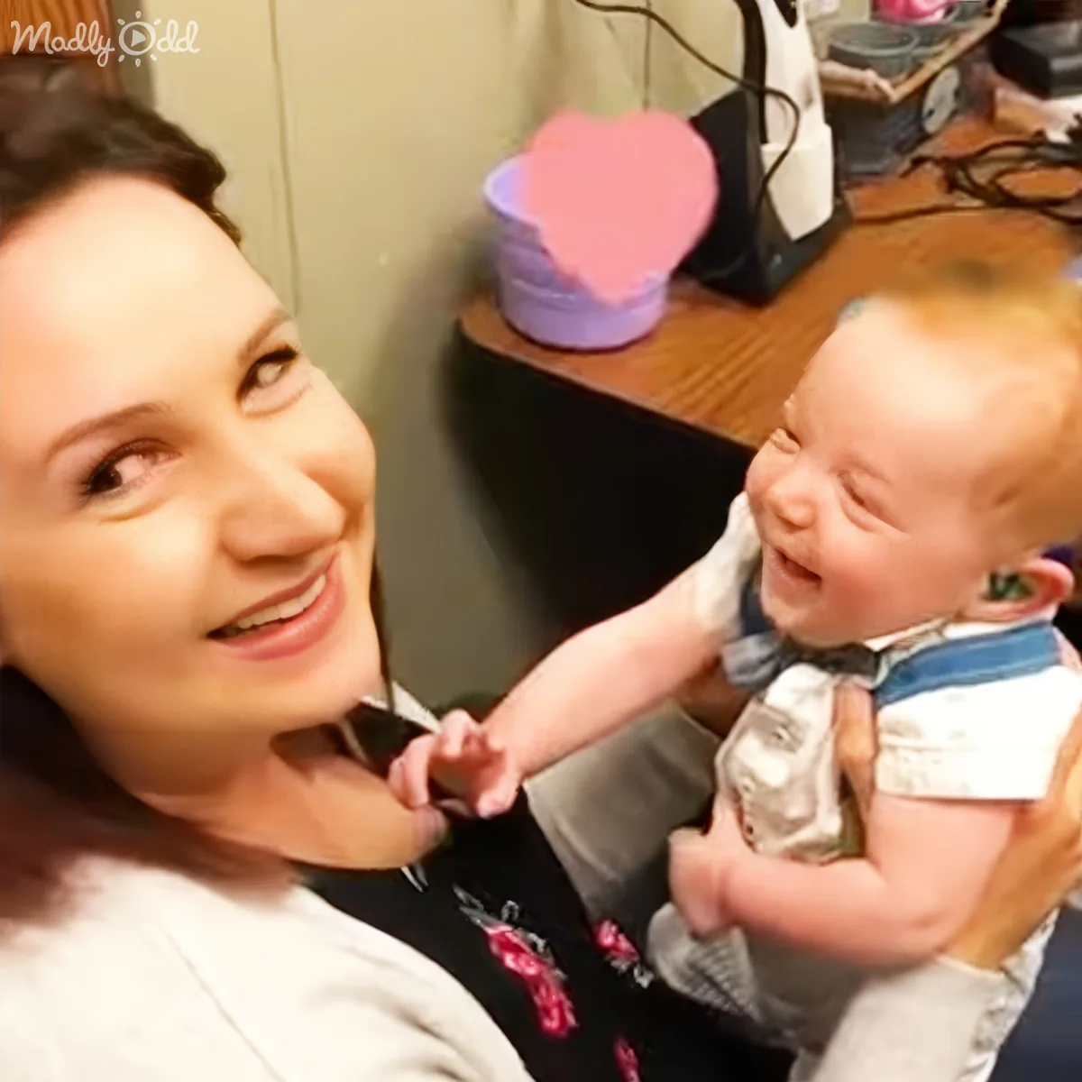 A woman holds a baby in a room with a heart-shaped object and a black appliance in the background.