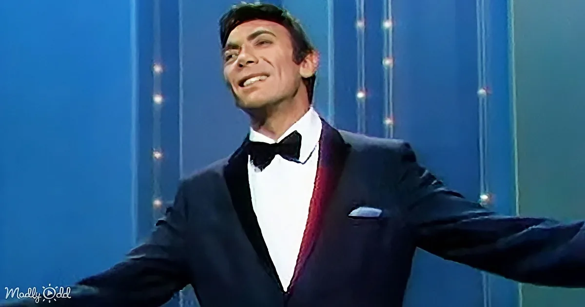 A man in a tuxedo with a bow tie and pocket square smiles with arms outstretched against a blue backdrop.