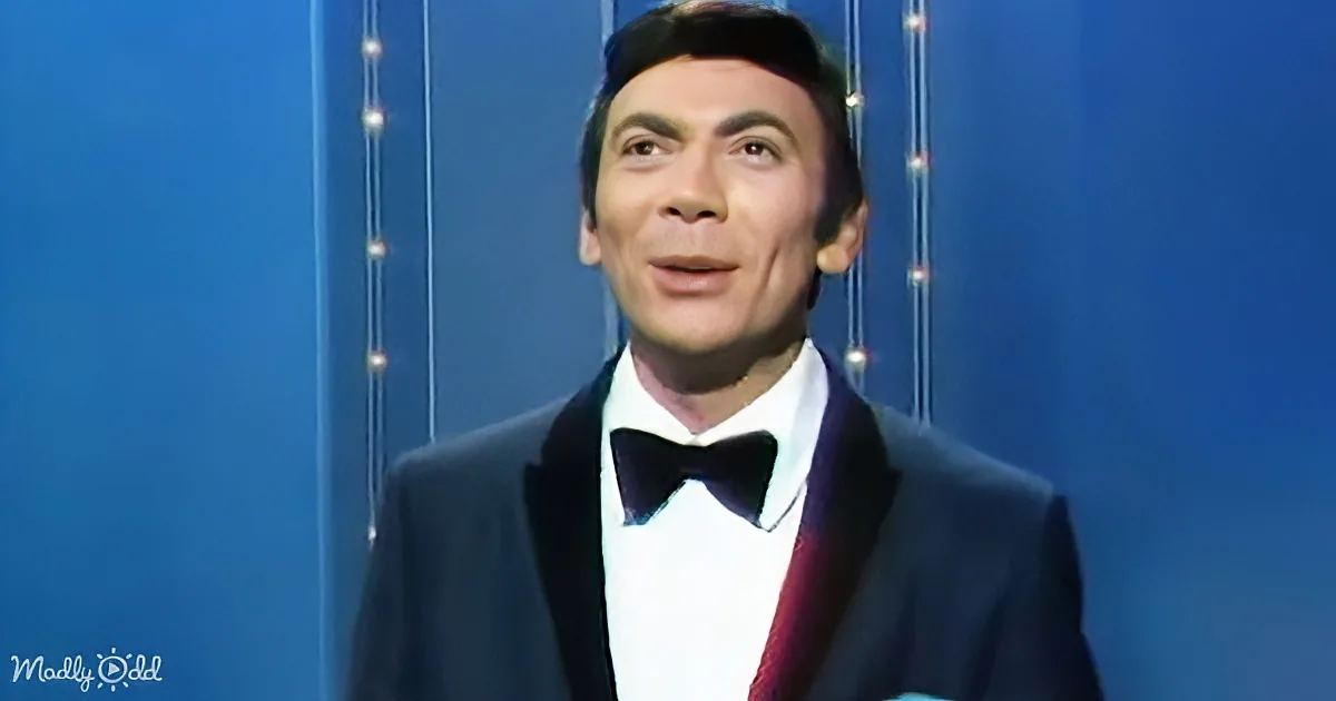 A man in a tuxedo with a bow tie stands against a blue backdrop with string lights.