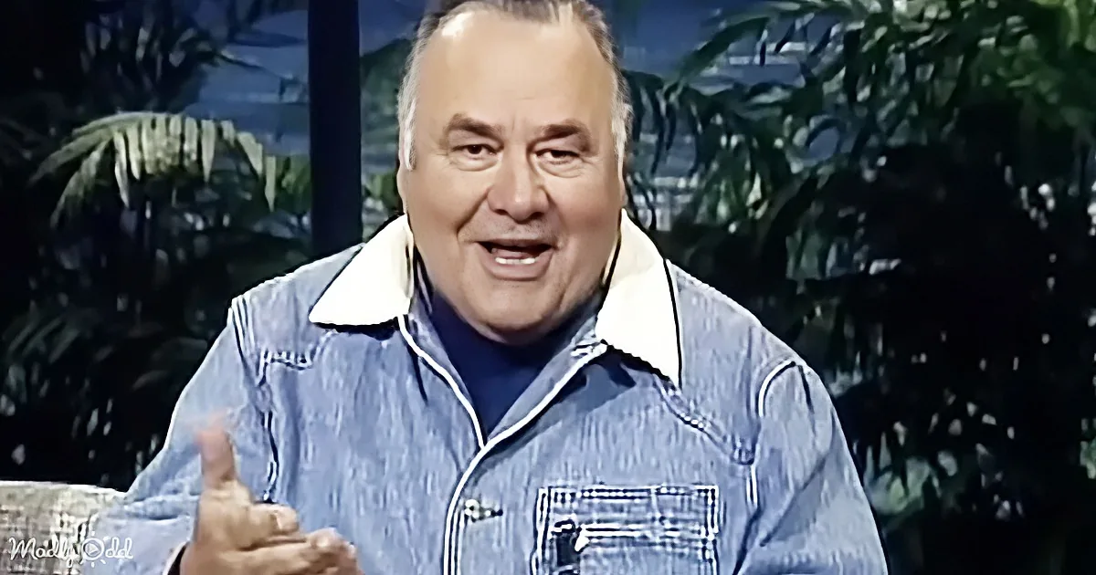 A man in a denim jacket gestures with his hand, smiling, against a tropical backdrop.