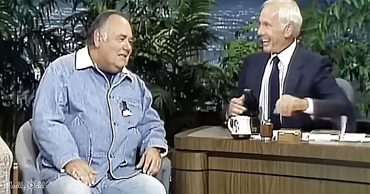 Two men are seated on a talk show set, one in a denim jacket, the other in a suit, both smiling and engaged in conversation.