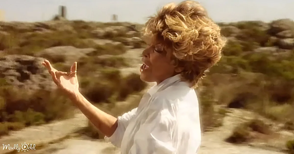 A person with short, curly hair in a white shirt stands outdoors, gesturing with their hand raised.