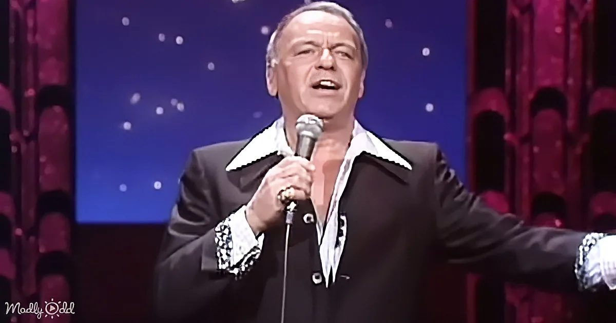 A man in a black suit with white accents sings into a microphone on stage.