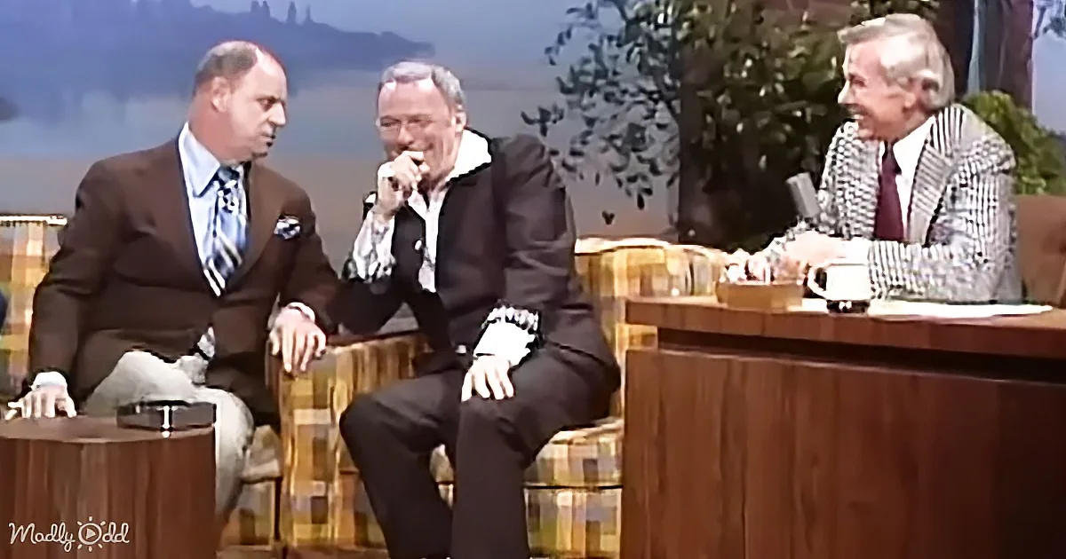 Three men in suits sit on a talk show set, one laughing, another gesturing, and the third holding a mug.