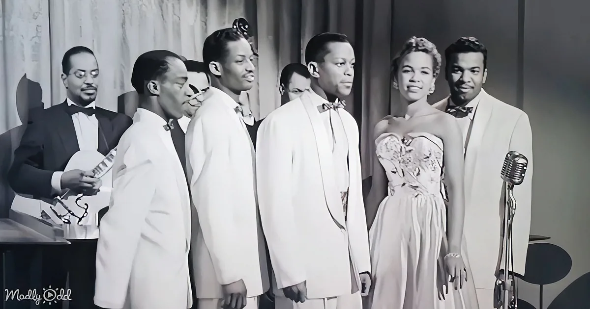 A group of six individuals in formal attire, including a woman in a dress, stands together on a stage, possibly performing.