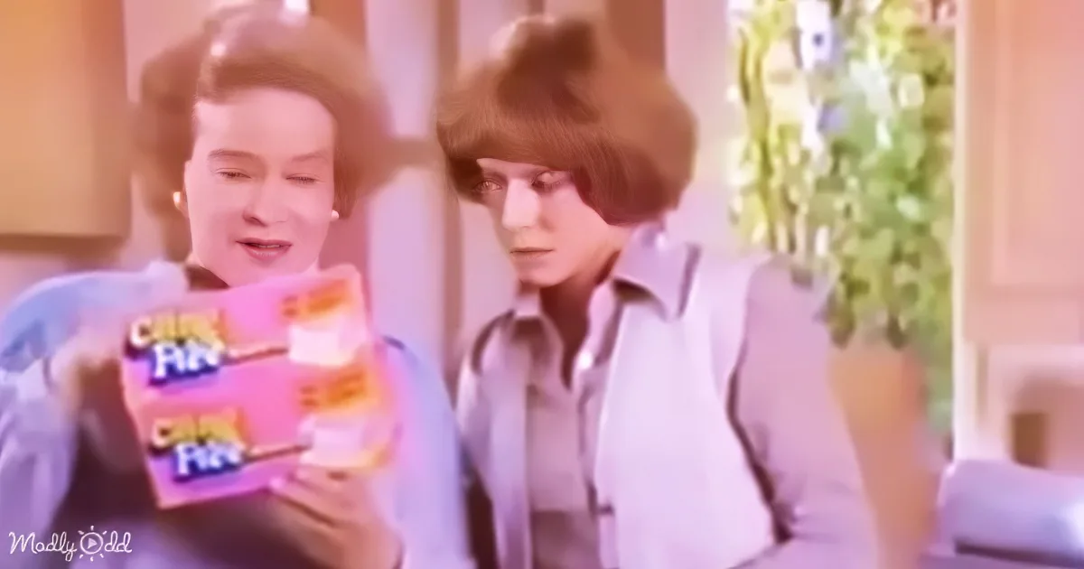 Two individuals are holding a pink cereal box, with one person wearing a light blue shirt and the other in a white shirt...
