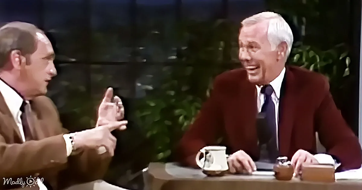 A man in a brown suit gestures animatedly while another man in a red suit listens and smiles.