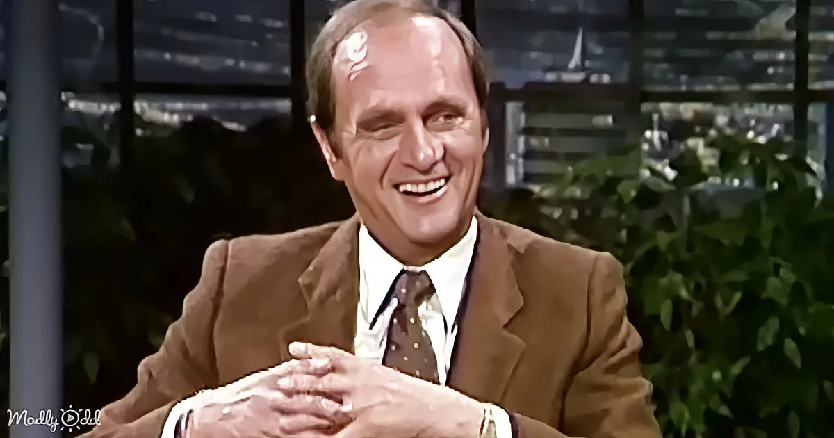 A man in a brown suit and tie smiles while clasping his hands, with a blurred background of greenery and city lights.