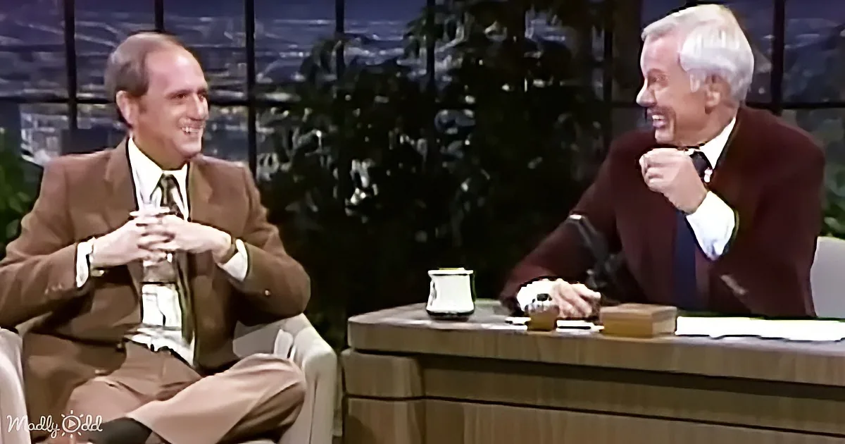 Two men in suits are seated, one laughing, on a talk show set with a cityscape background.
