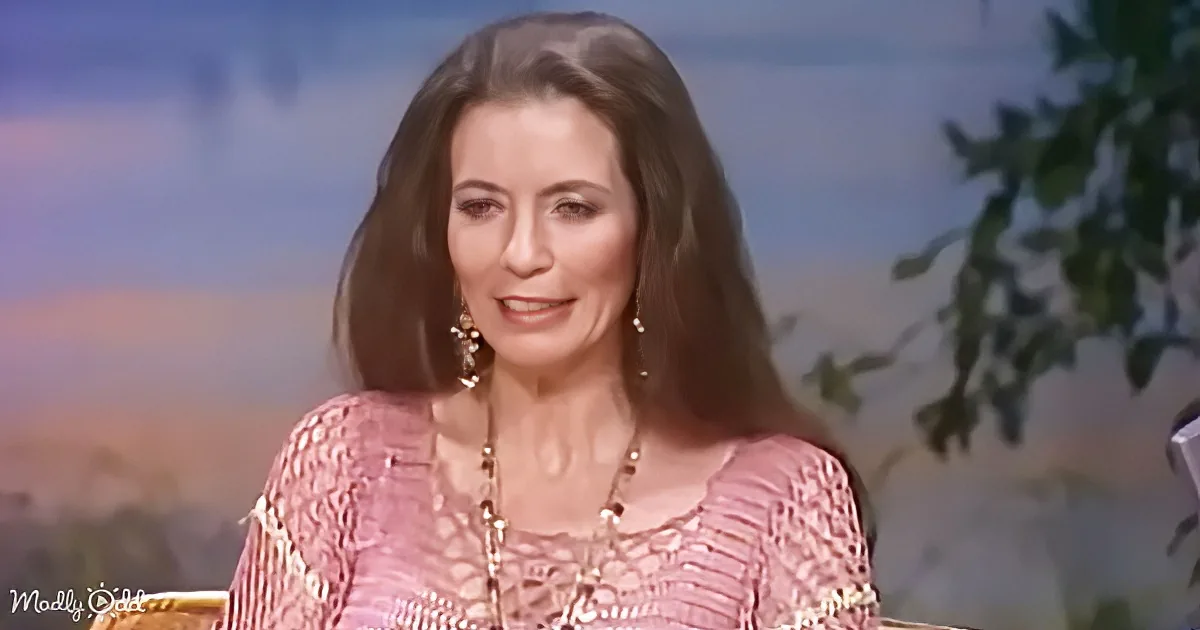 A woman with long brown hair, wearing a pink patterned top and a necklace, is seated in front of a scenic backdrop.