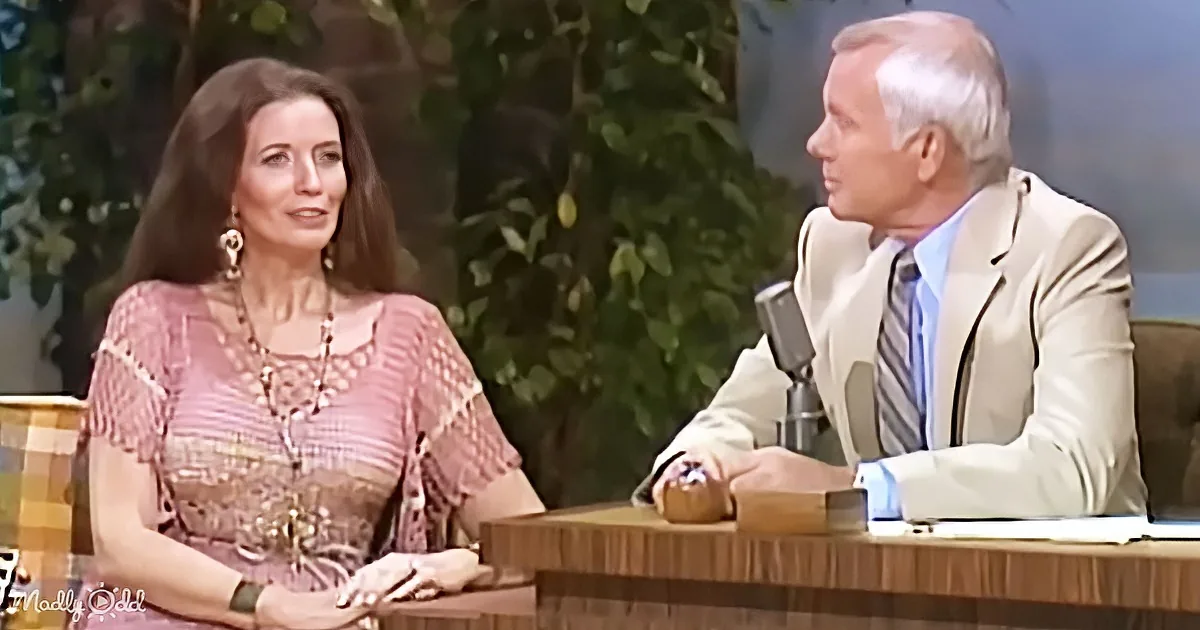 Woman in pink dress and necklace sits at table, conversing with man in suit and tie.