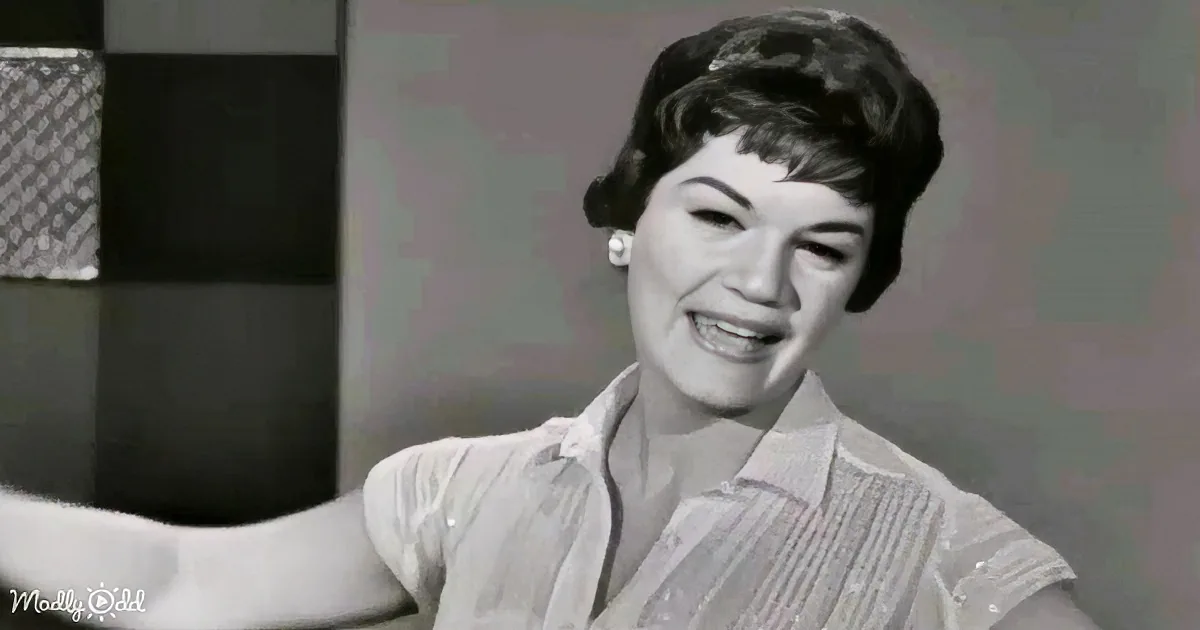 A smiling woman in a striped shirt extends her arm, wearing earrings, in a black-and-white setting.