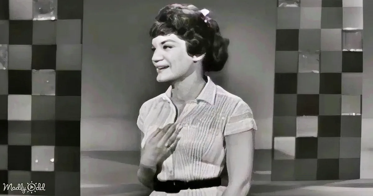 A woman in a striped shirt and belt stands against a checkered background, smiling.