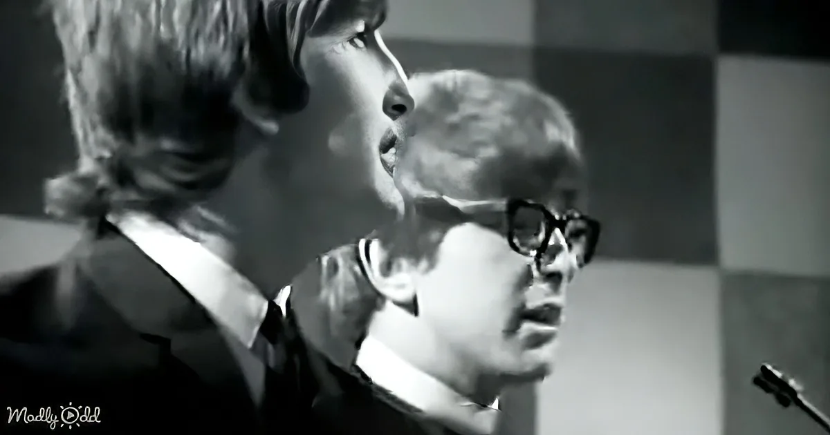 A man with glasses and a microphone, possibly singing or speaking, with another person nearby.