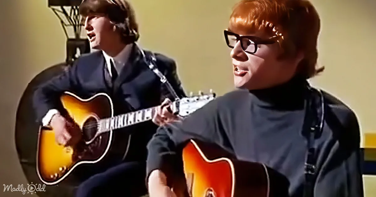 A person with red hair and glasses plays guitar, another person in a suit sings behind.