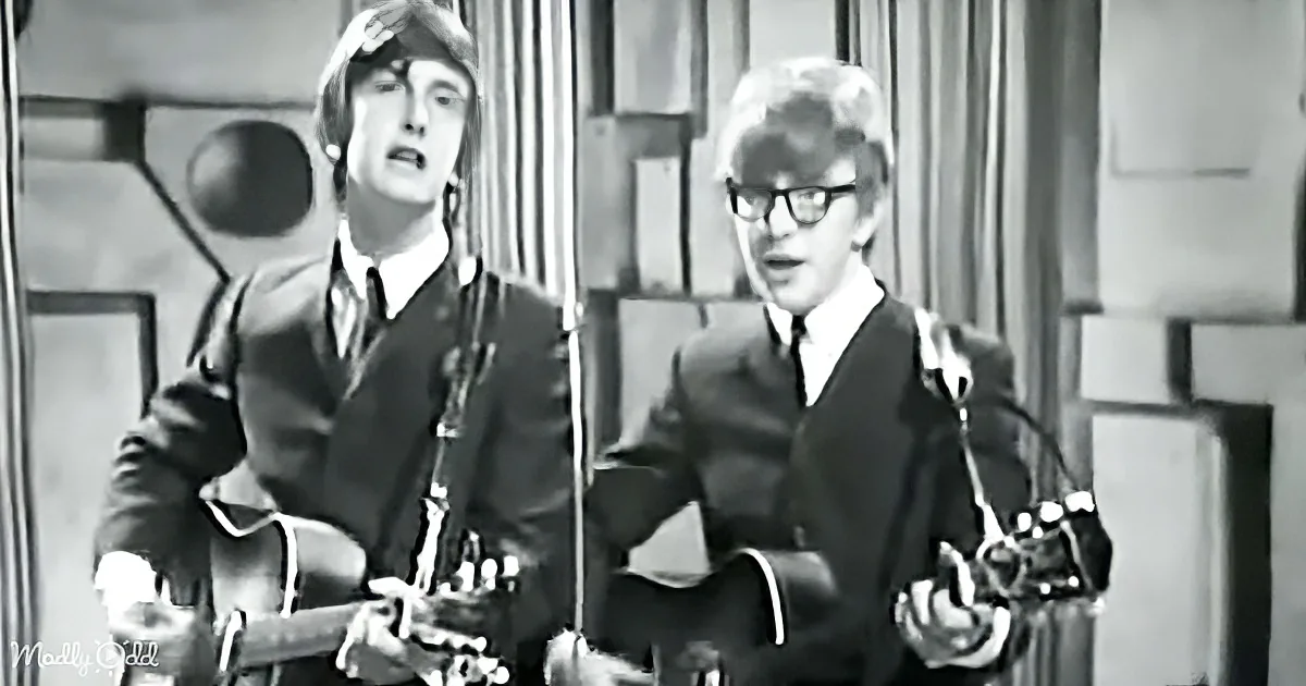 Two musicians in suits play guitars, one singing, in a black-and-white setting.