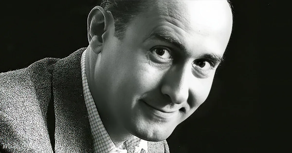 A man with a bald head, wearing a textured jacket and plaid shirt, smiles slightly in a black-and-white portrait.