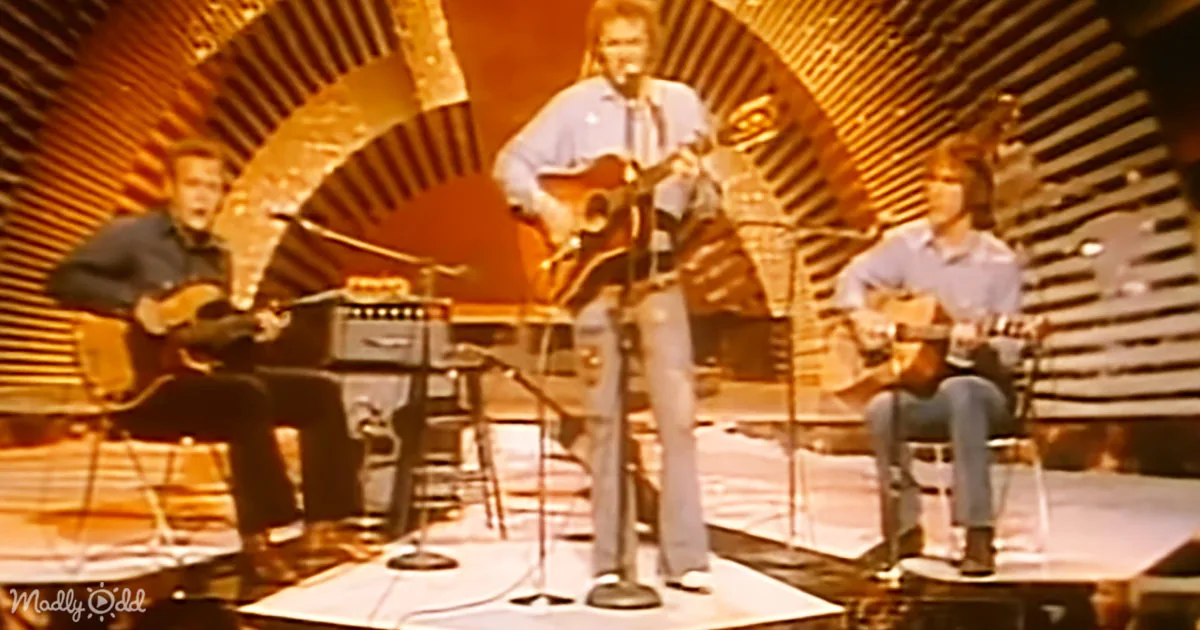 A man in a blue shirt and jeans stands center stage with a guitar, singing into a microphone.