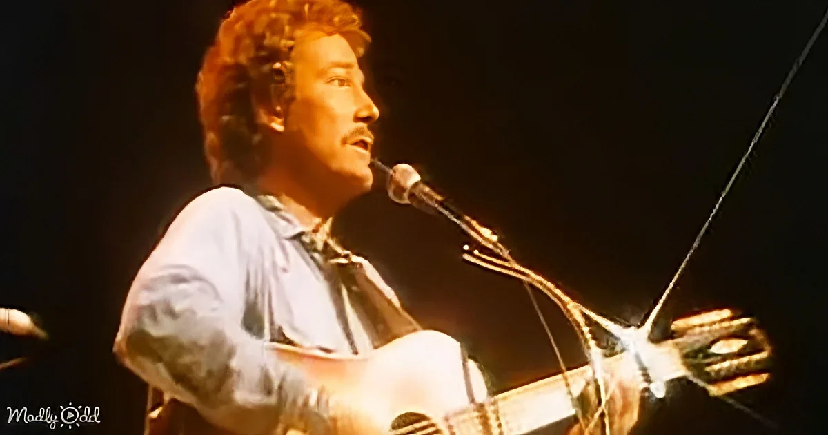 A man with curly hair plays an acoustic guitar and sings into a microphone on stage.