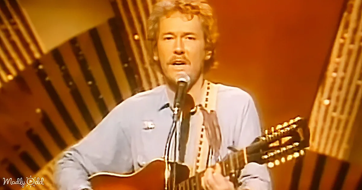 A man with curly hair plays an acoustic guitar and sings into a microphone.