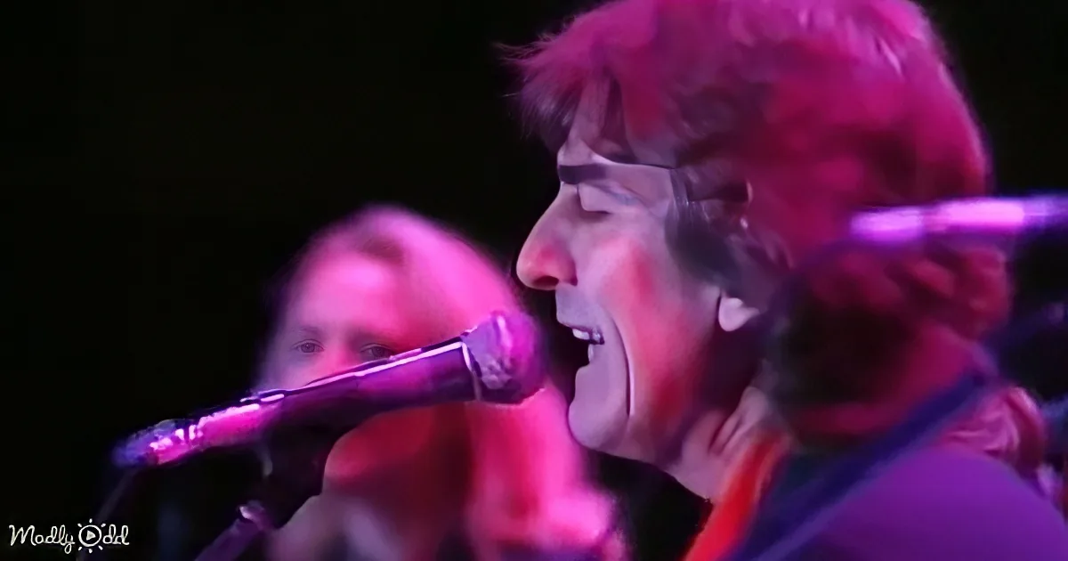 A person with curly hair sings into a microphone under vibrant purple stage lighting.