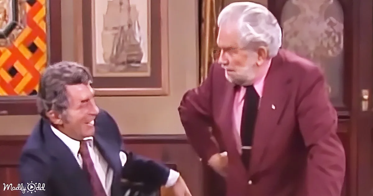 A man in a maroon suit is leaning forward, engaging in conversation with another man in a dark suit, both in a warmly lit...