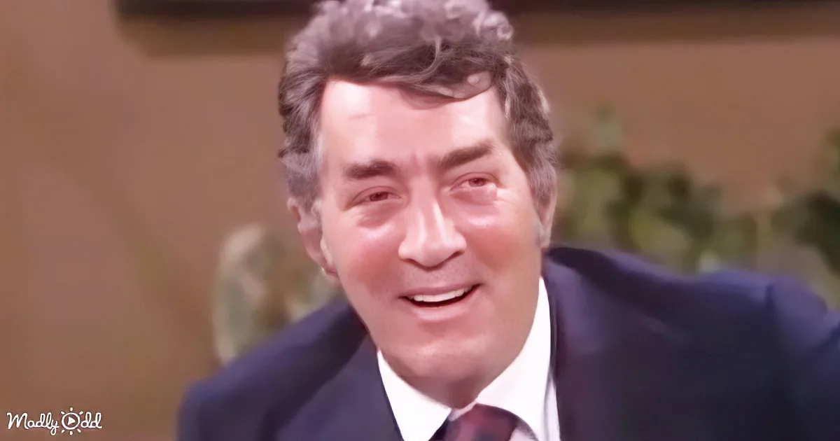 A man in a suit and tie smiles warmly, with a blurred background and a plant visible.