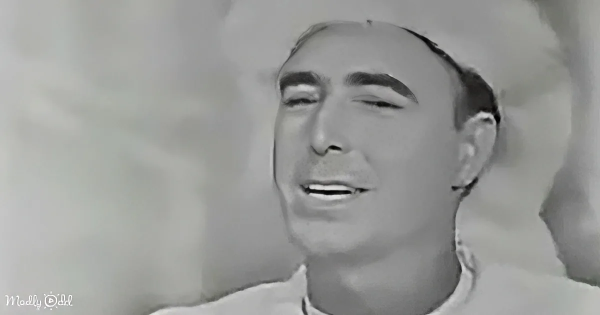 A man with a receding hairline and a white shirt is shown in a close-up, appearing to be singing or speaking.