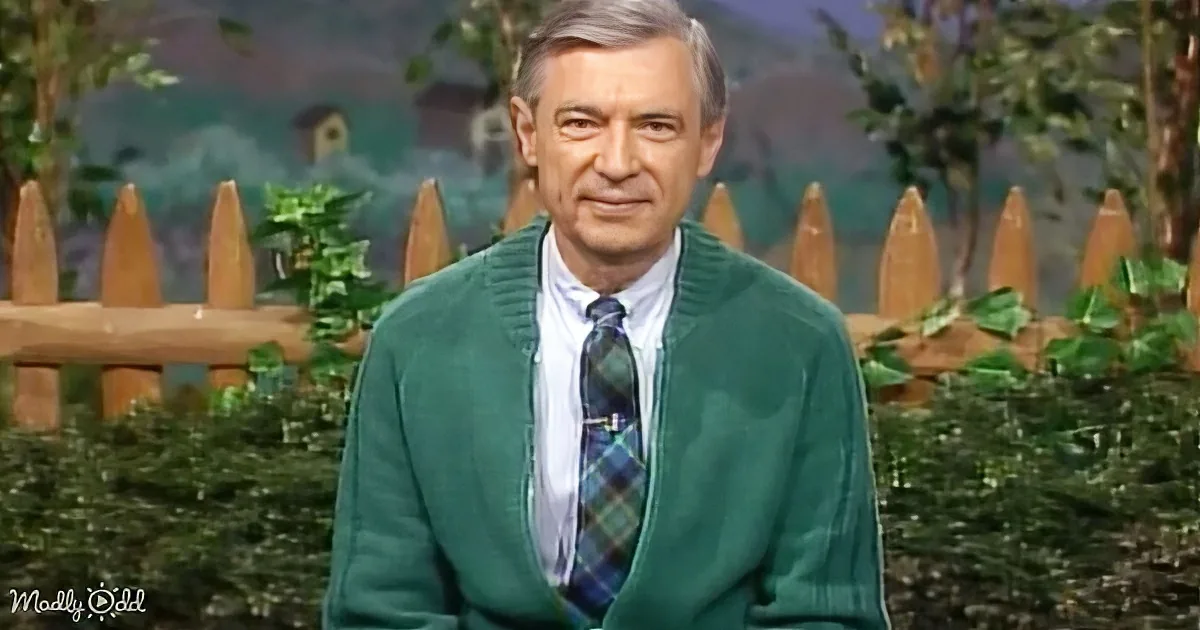 A man in a green sweater and plaid tie stands in front of a wooden fence and greenery backdrop.