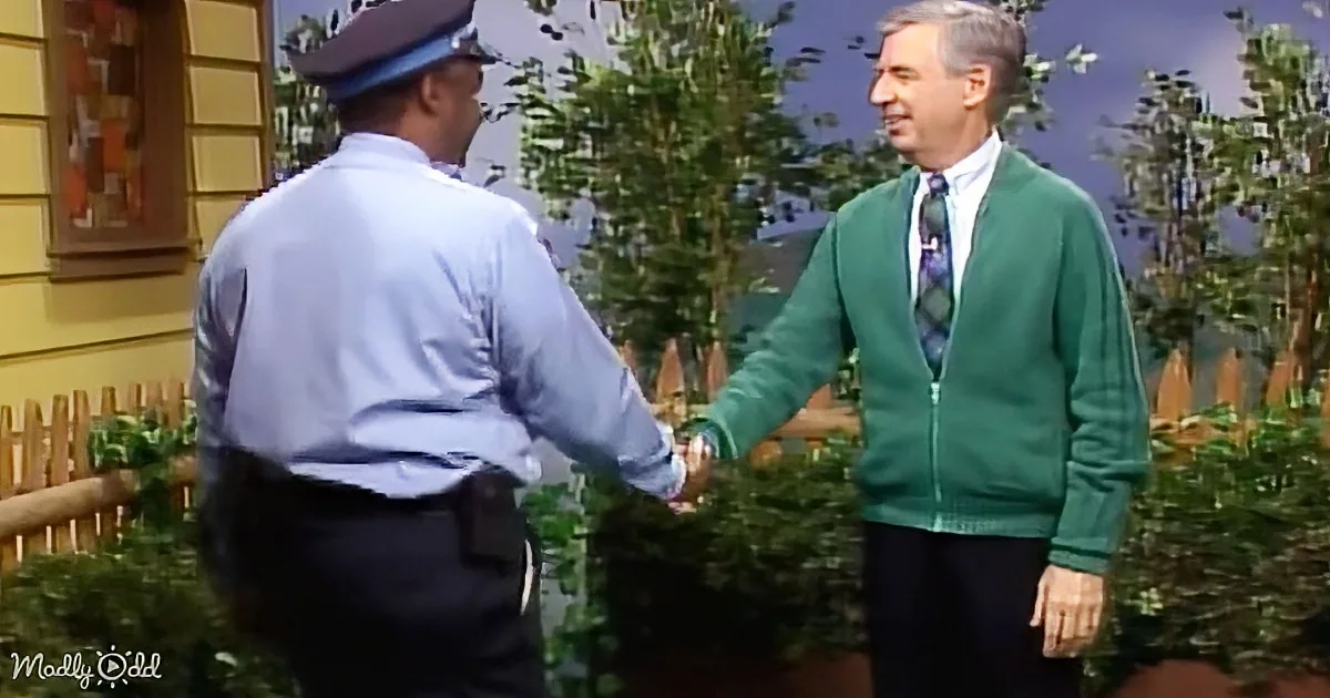 A man in a green sweater and tie shakes hands with a uniformed individual in a blue shirt and hat.