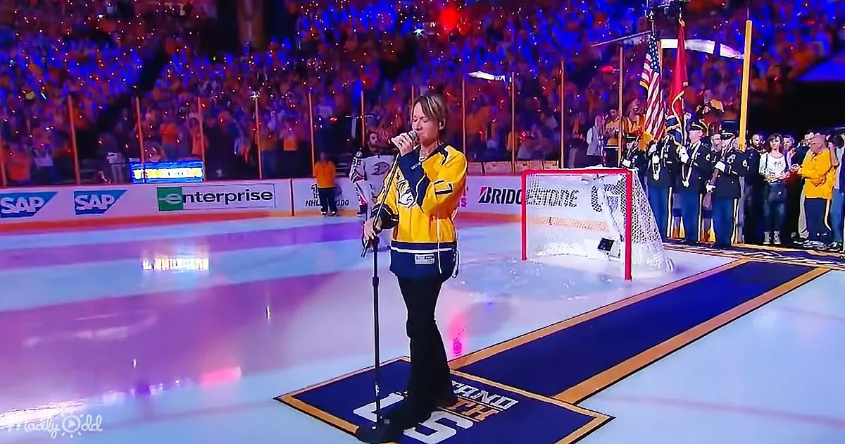A person in a yellow jacket sings into a microphone on an ice rink, with a crowd and military personnel in the background.