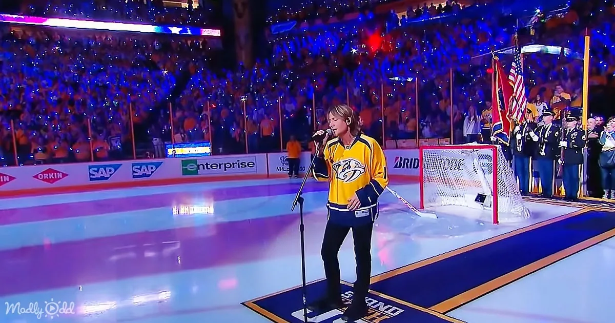 A person in a yellow jersey sings into a microphone on an ice rink with a crowd and flags in the background.