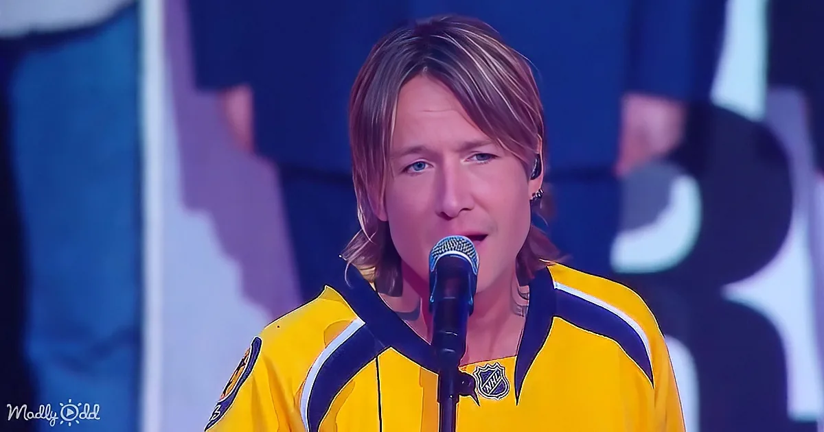 A person in a yellow jersey sings into a microphone, wearing an earpiece, with a blue background and a logo visible.