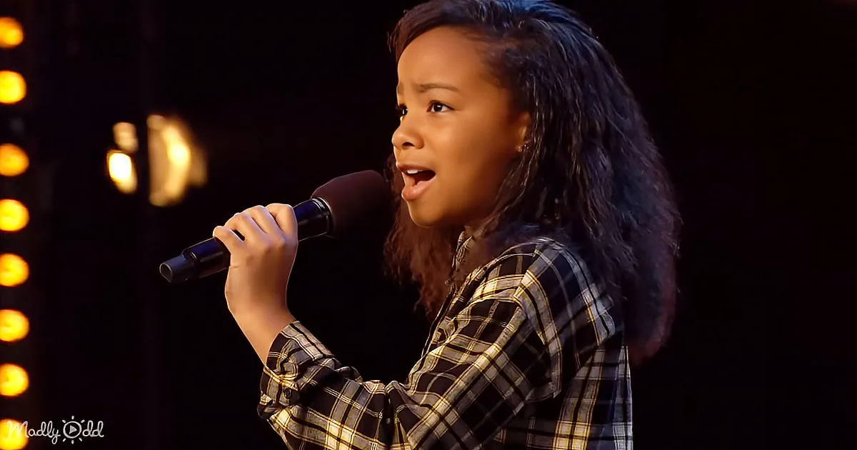 A young child in a plaid shirt sings into a microphone on stage, with warm lighting in the background.