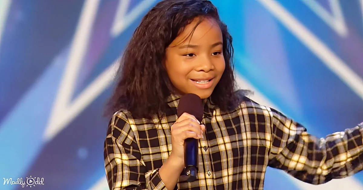 A young girl in a plaid shirt holds a microphone, smiling, with a colorful background.