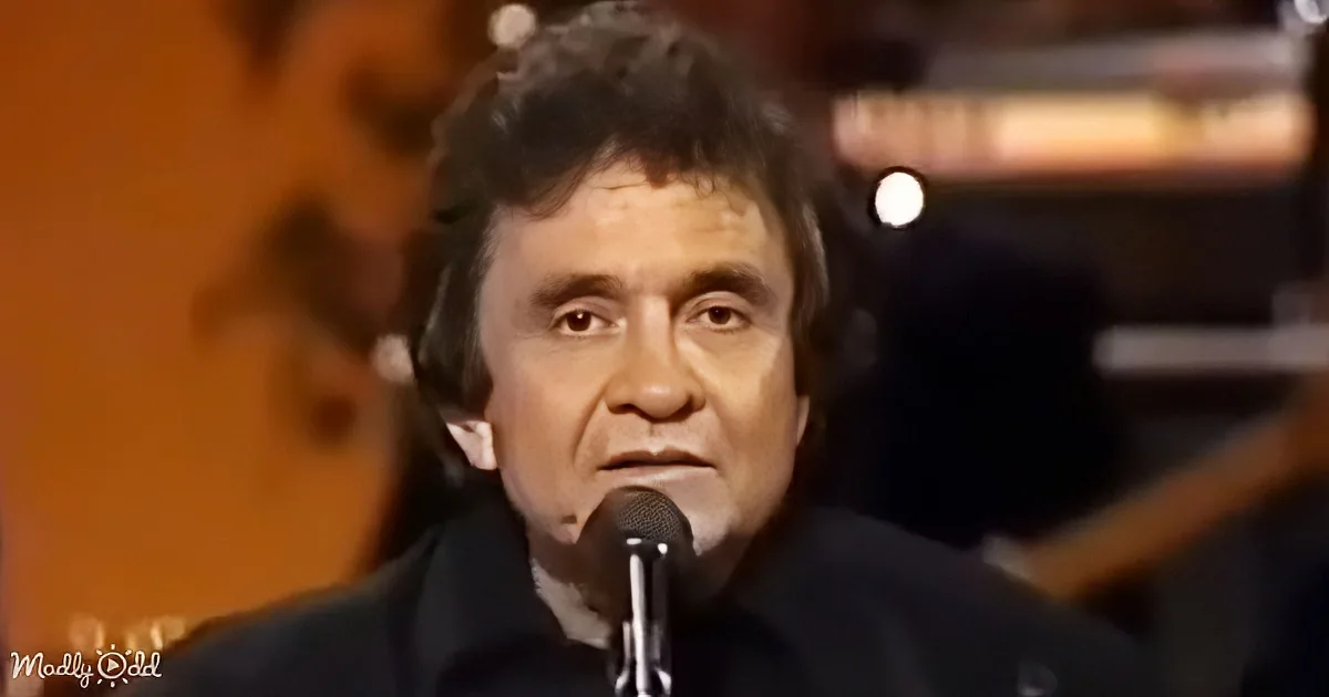 A man with dark hair and a black suit sings into a microphone, with a blurred background of musical instruments.