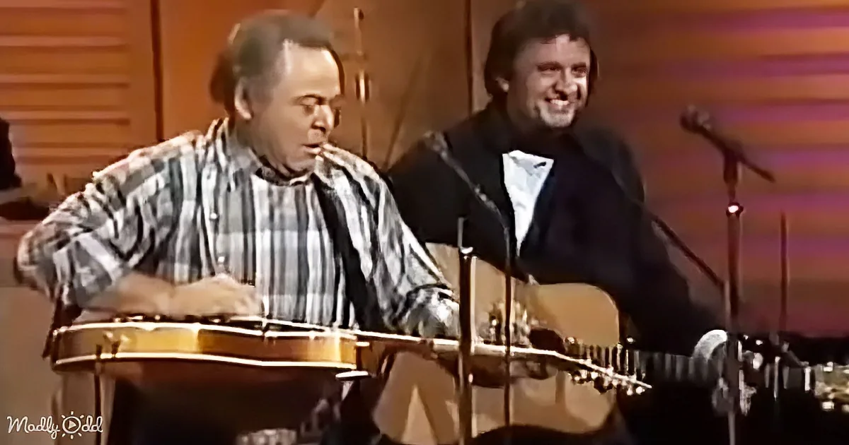 A man in a plaid shirt plays a banjo while another man in a suit smiles nearby.