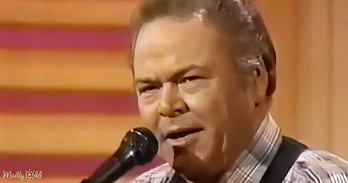 A man in a plaid shirt sings into a microphone, wearing an earpiece, with a colorful background.