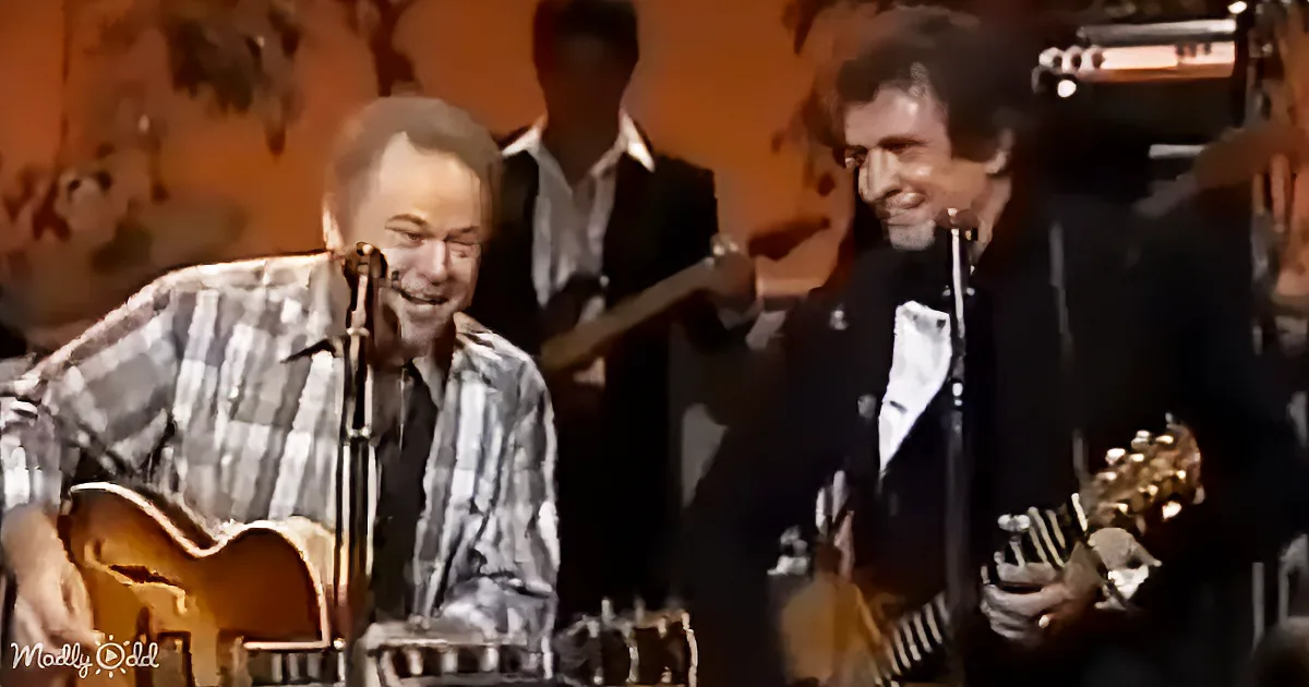 Two smiling musicians play guitars on stage, one in plaid shirt, another in dark suit, with a band in the background.