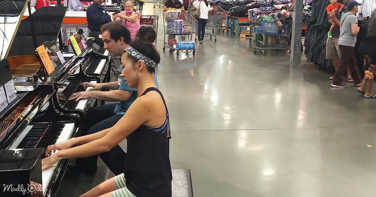 A woman and man play a grand piano in a store aisle, surrounded by shoppers and shopping carts.