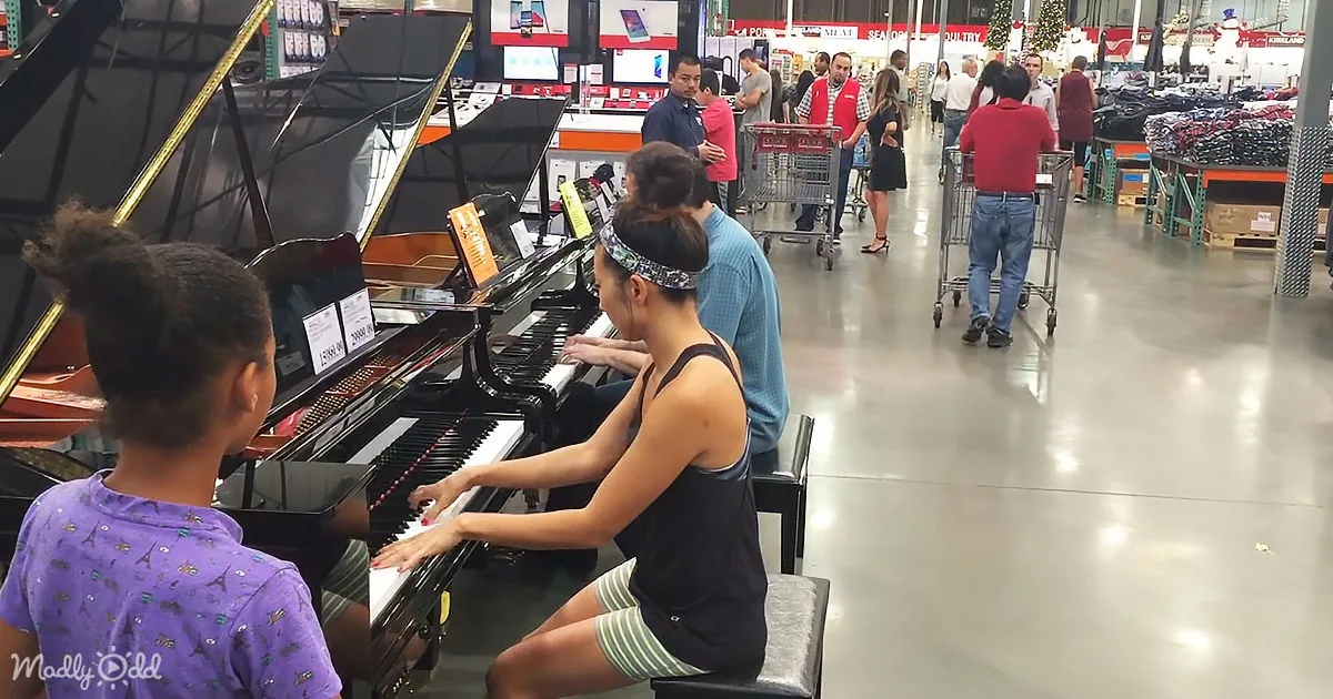 A woman plays a piano in a store aisle, with a child watching, while shoppers browse in the background.