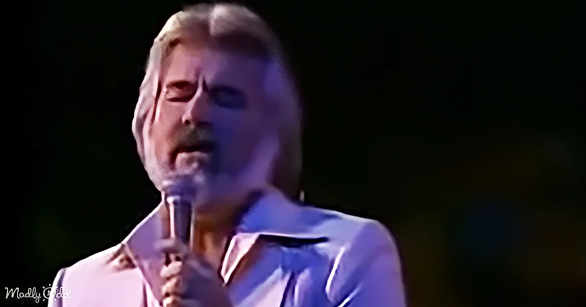 A man with closed eyes holds a microphone, wearing a white shirt, against a dark background.