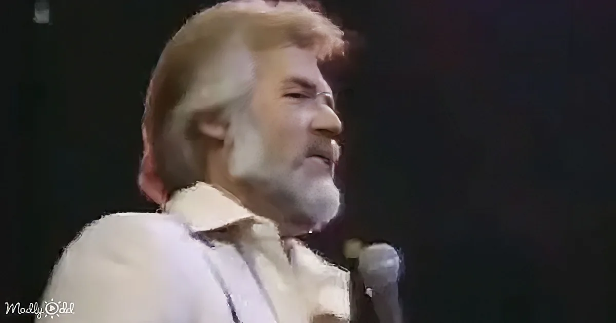 A man with white hair and beard, wearing a white shirt, sings into a microphone on stage.