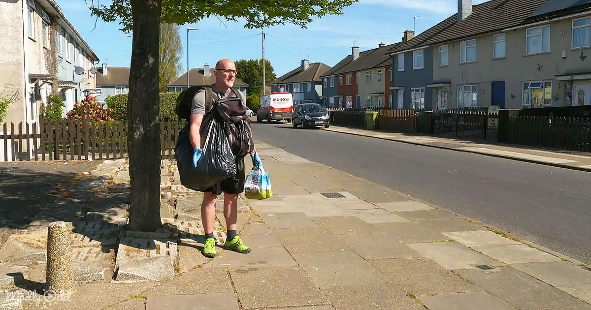 A man stands on a sidewalk carrying a trash bag and groceries, wearing gloves and a backpack.