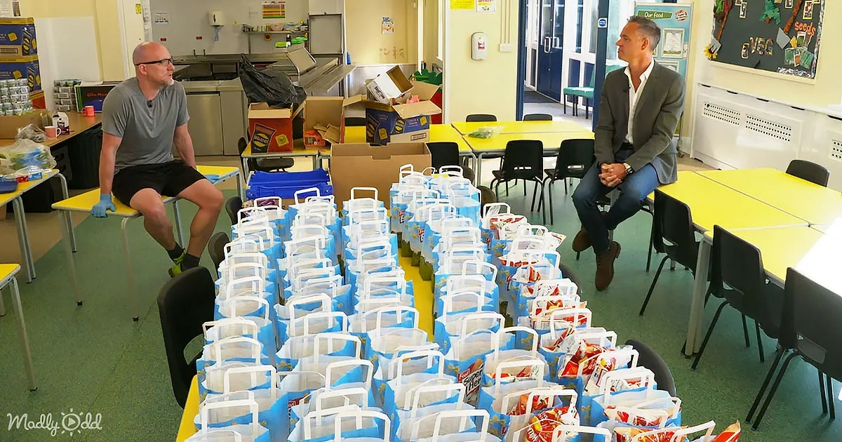 Two men sit in a room with yellow tables, surrounded by blue and white bags filled with food.