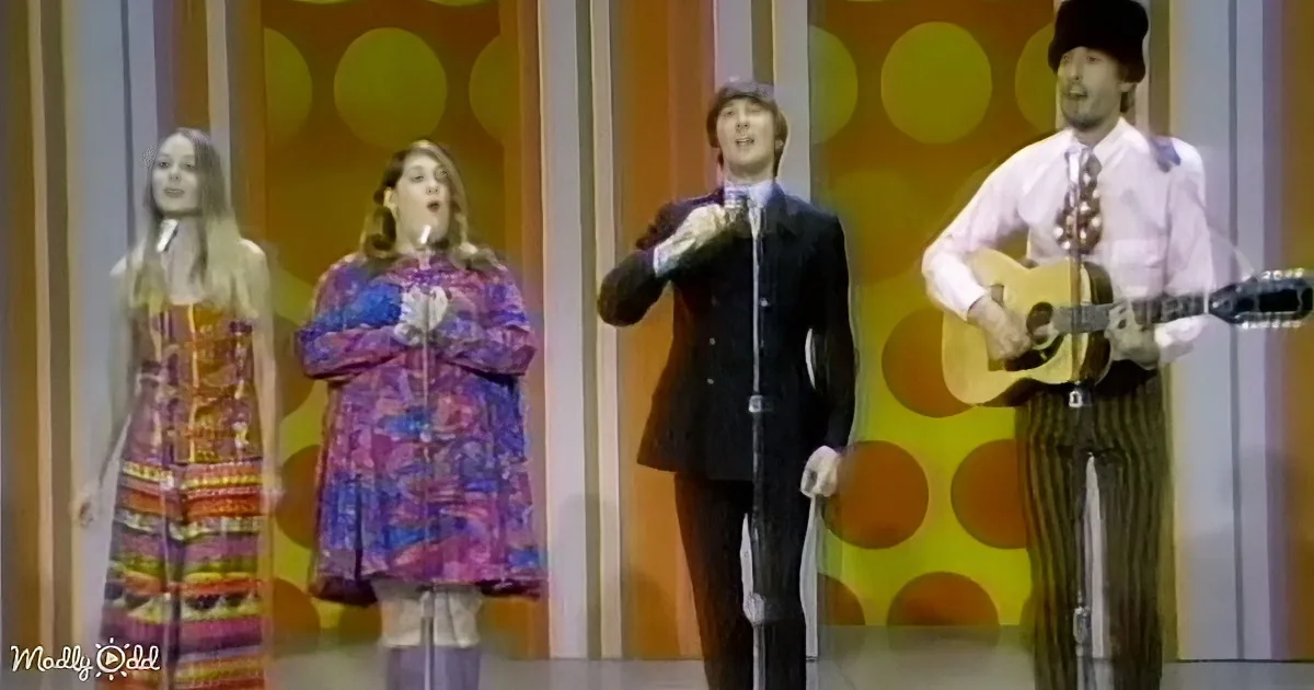 A man in a suit sings into a microphone, accompanied by two women and a guitarist on a stage with a colorful background.