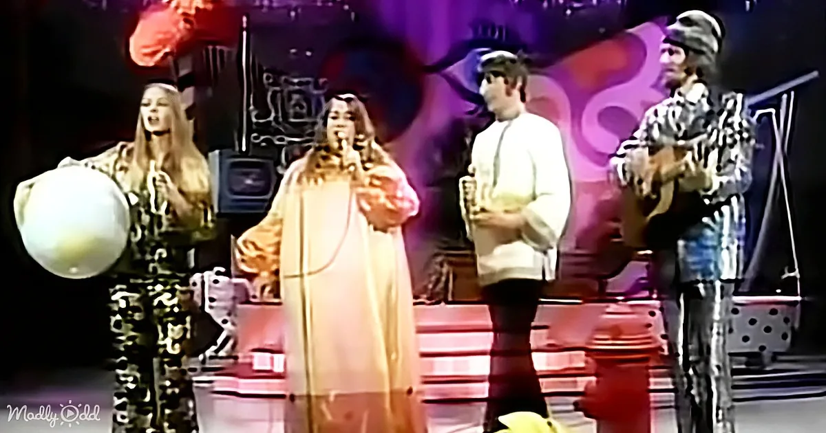 A woman in a long white dress sings on stage with a band, including a guitarist and a drummer.