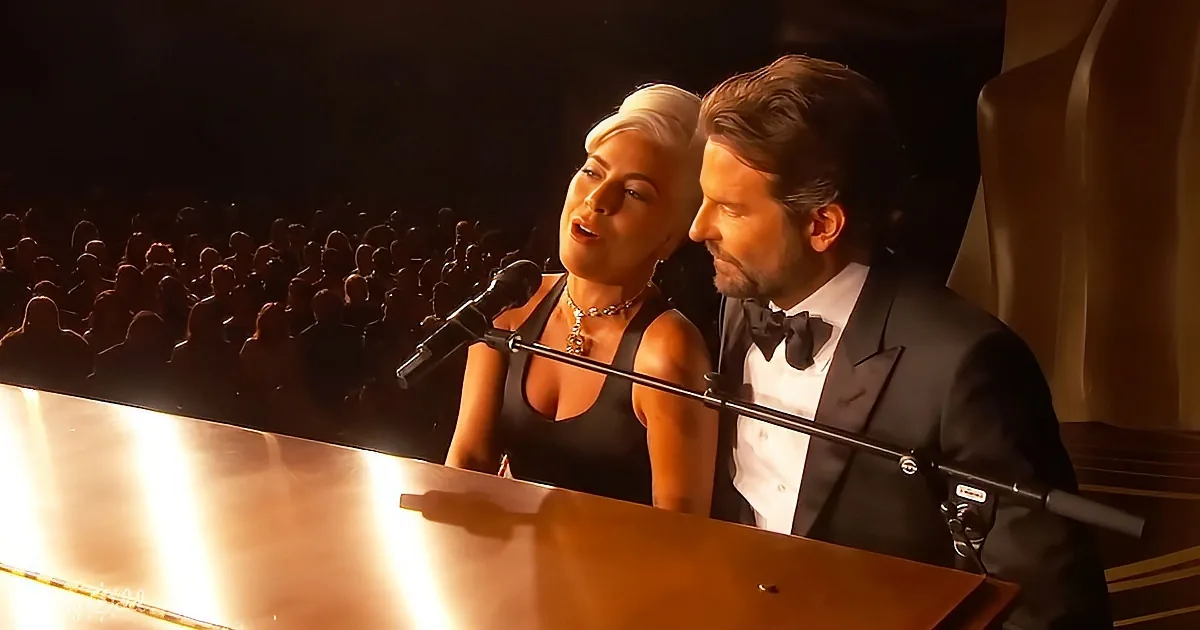 A man and woman sing at a piano on stage, with a crowd in the background.