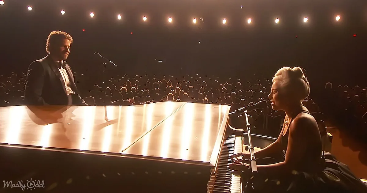 A woman sings at a piano while a man stands beside her on stage in front of an audience.
