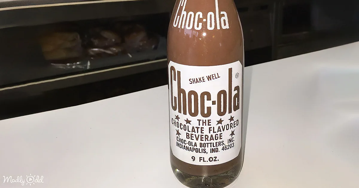 A bottle of Choc-ola, a chocolate-flavored beverage, sits on a white surface with a glass and donuts in the background.